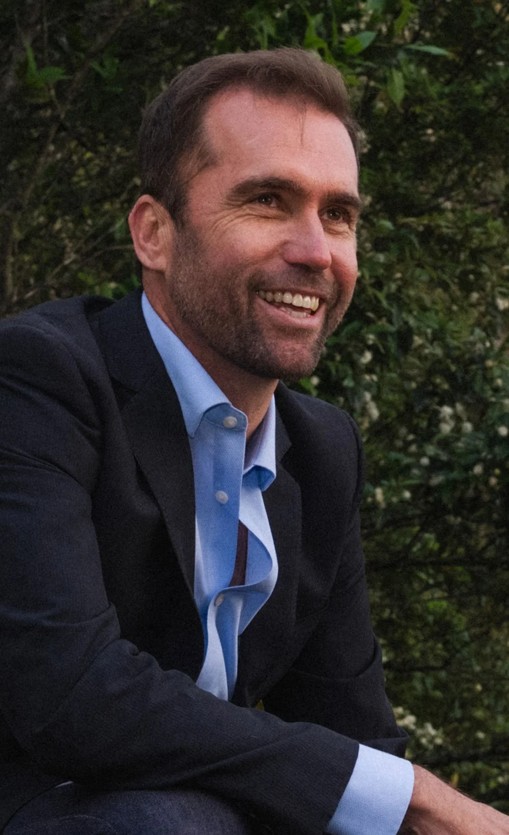 Brad McGee smiling in a suit and light blue shirt outdoors surrounded by green foliage.