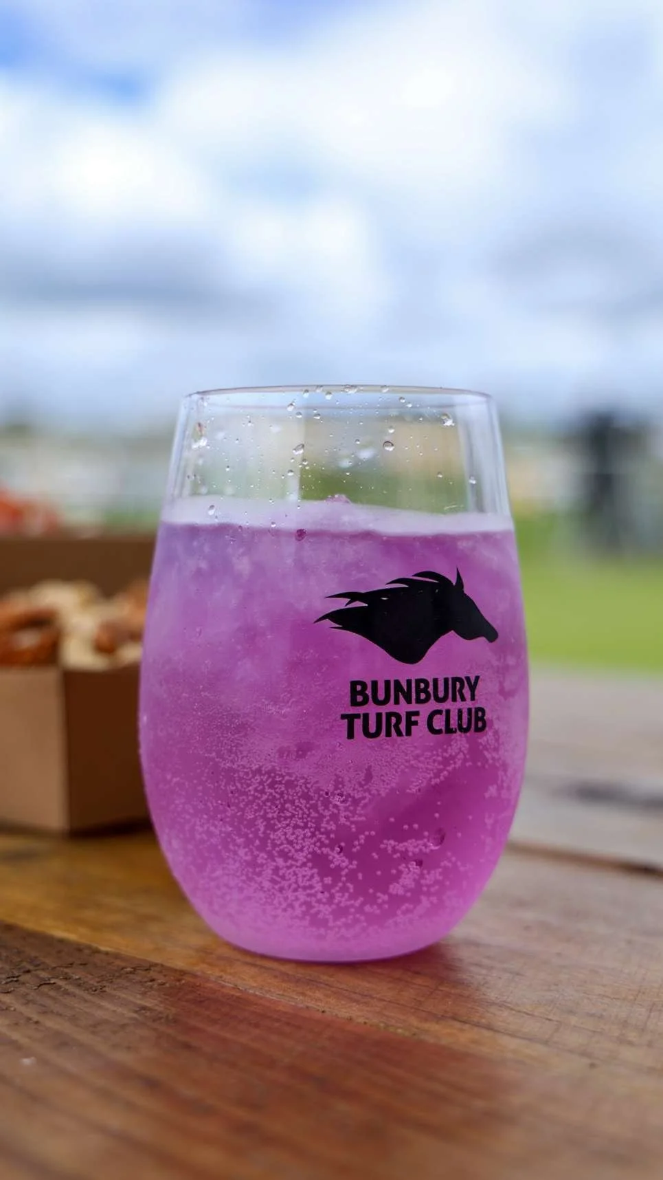 A clear glass with purple sparkling beverage and bubbles, featuring the Bunbury Turf Club logo with a horse's head silhouette, set on a wooden surface outdoors.