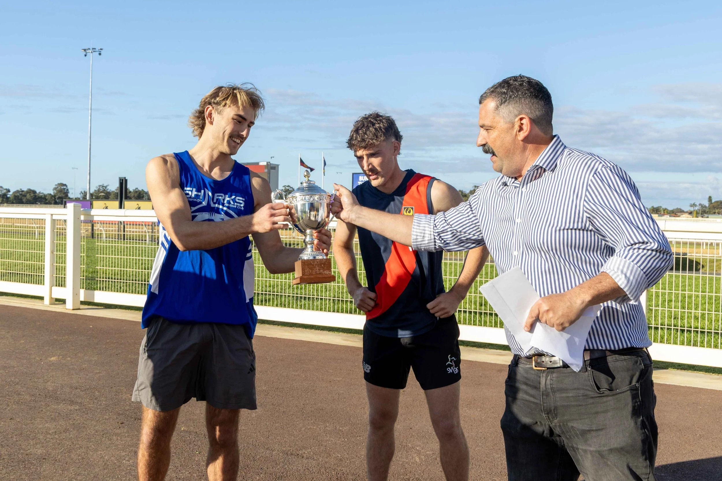 A man in a striped shirt handing a trophy to a young athlete in a blue sports jersey, while another athlete in a black and red sports outfit looks on, on an outdoor running track.