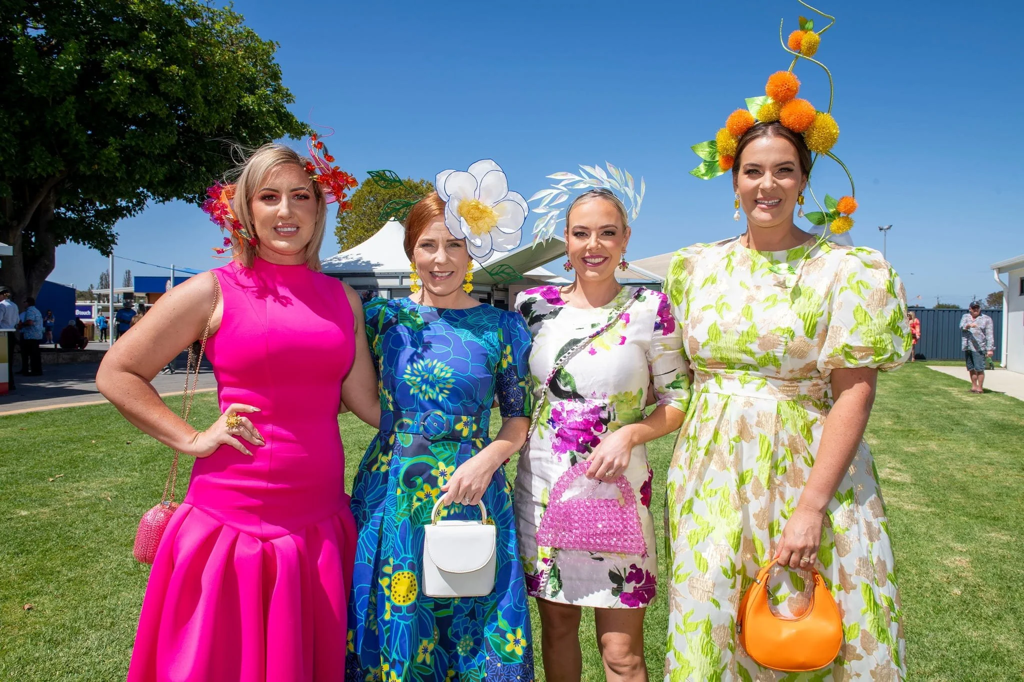 Four women standing outdoors on grass, dressed up in colorful, floral, and vibrant outfits with elaborate floral headpieces, smiling for the camera at a sunny event.