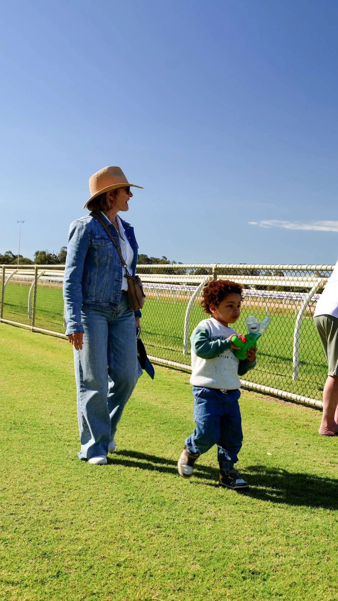 A woman wearing a wide-brimmed hat and denim jacket stands at the Bunbury Turf Club, looking at a young child holding a green toy and a plastic cup.