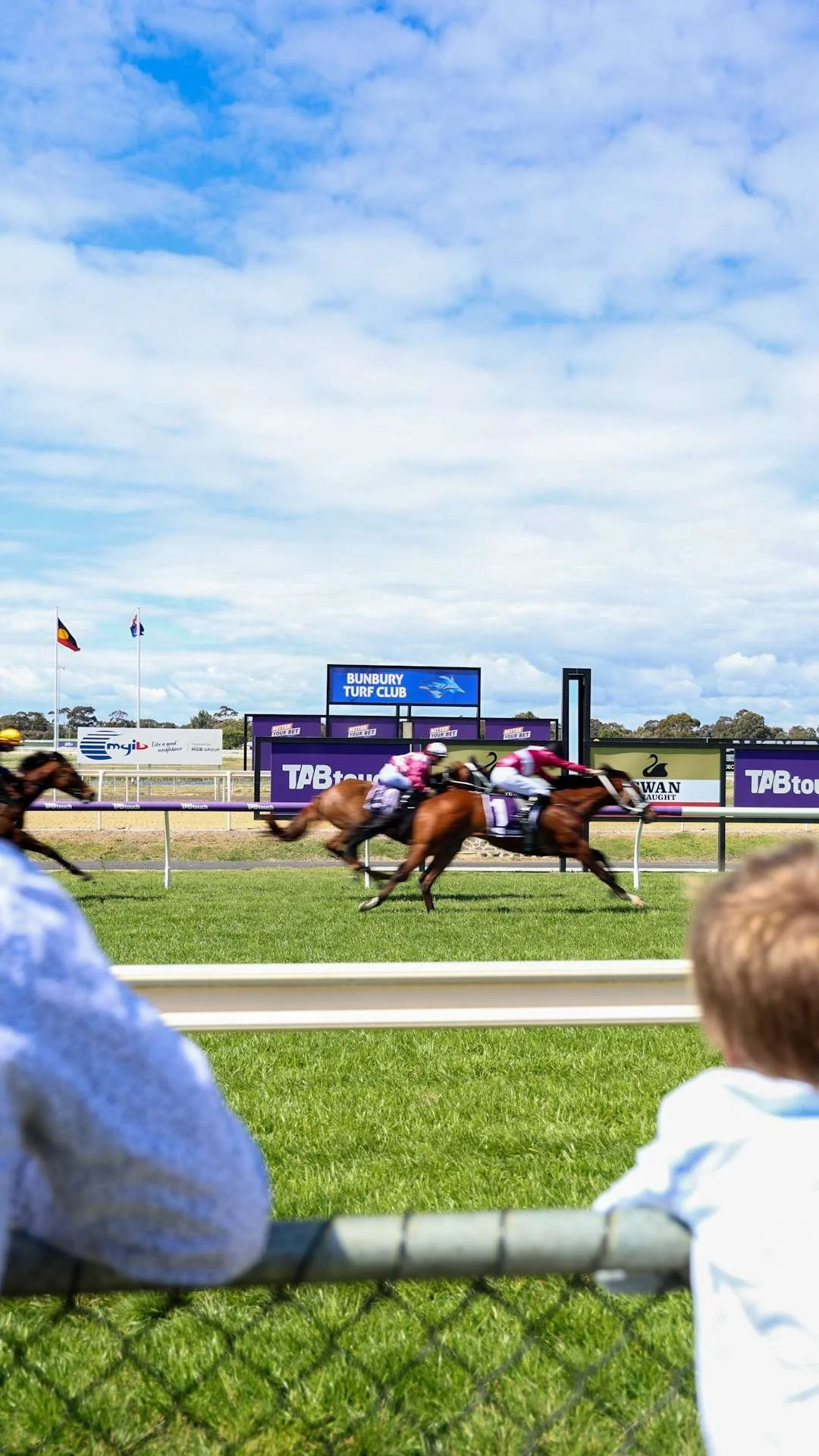 Horse race at Bungbury Turf Club, with several horses and jockeys racing on the track under a partly cloudy sky. Spectators are watching from behind the fence.