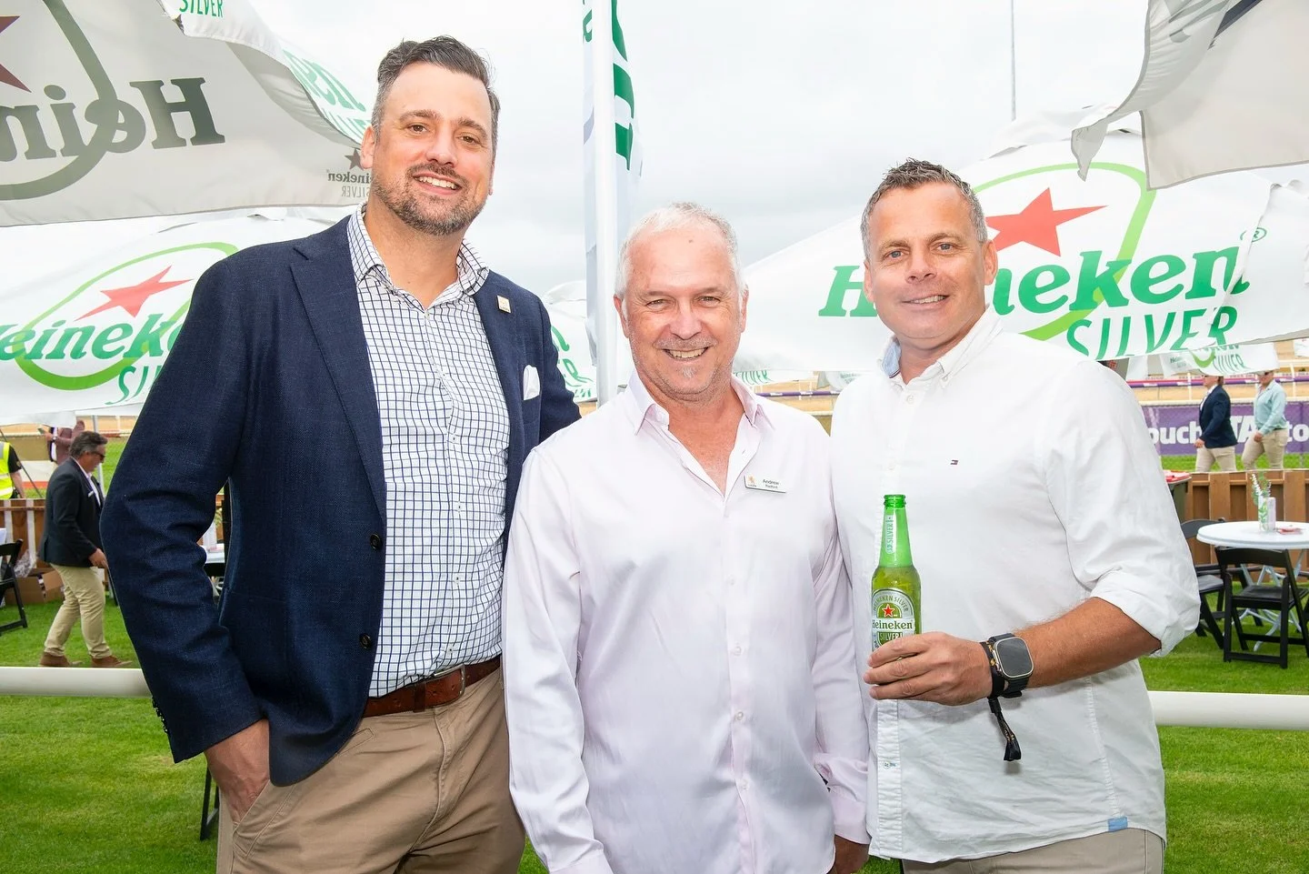 Three men smiling at an outdoor event, with Heineken Silver umbrellas in the background, one holding a Heineken beer bottle, dressed in business casual attire