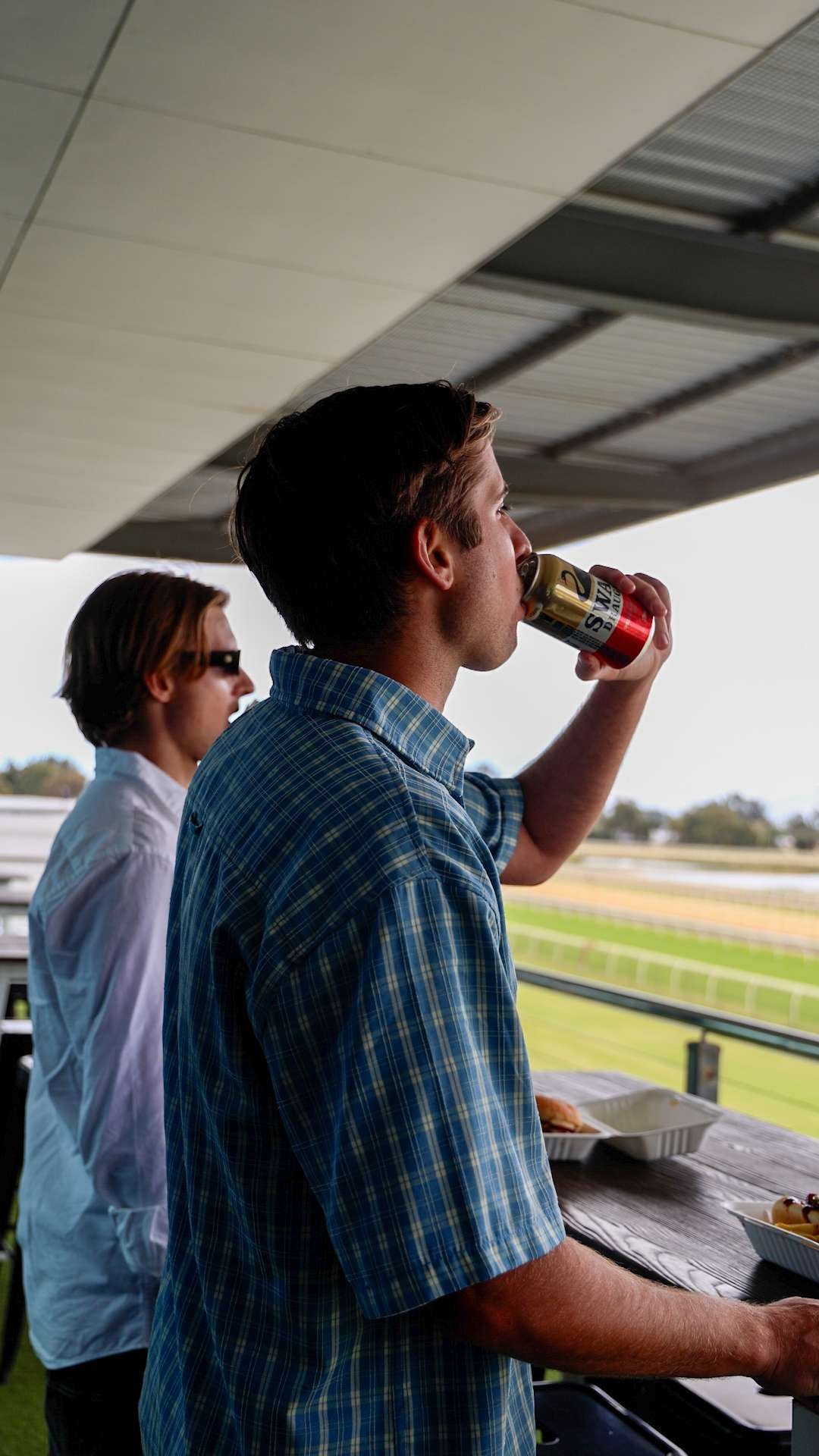 Two men at a racetrack, one drinking a soda and the other looking toward the race track, with food containers on the table in front of them.