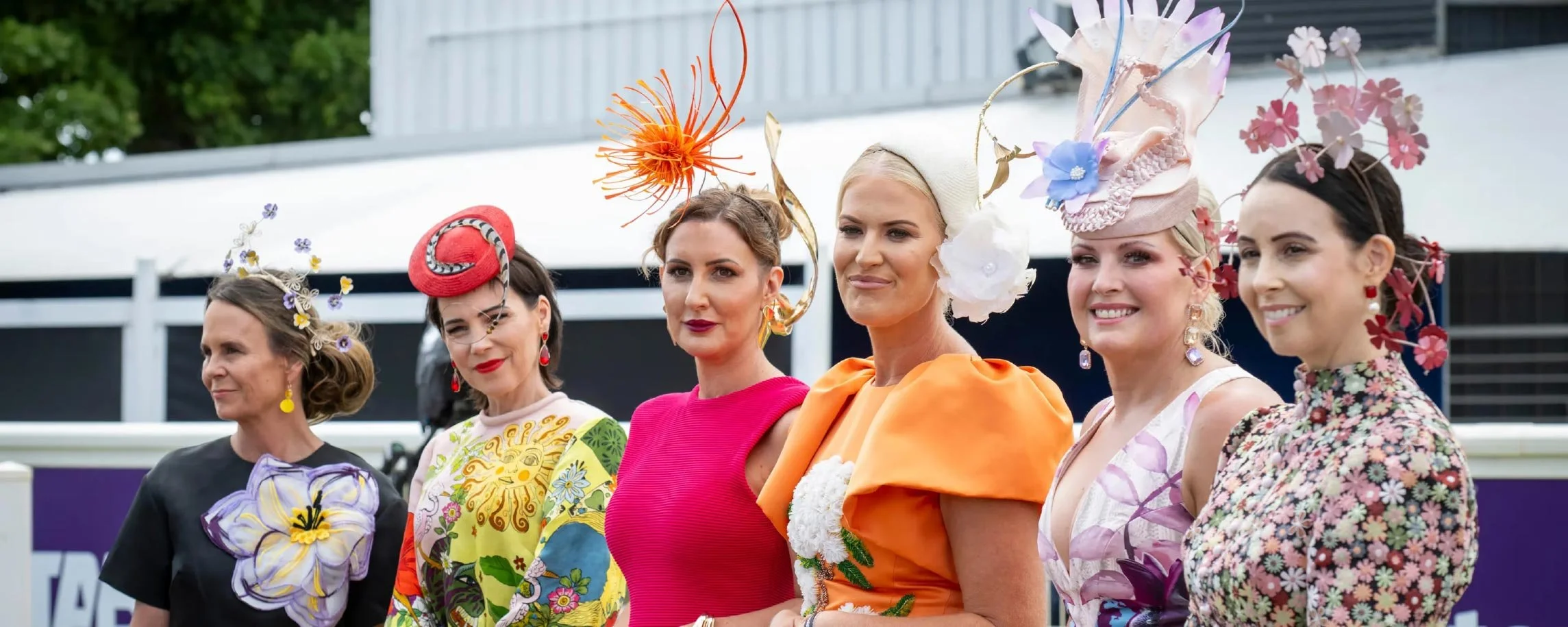 Six women at a race day event wearing colourful, floral, and uniquely designed hats and outfits, being judged for fashion on the field