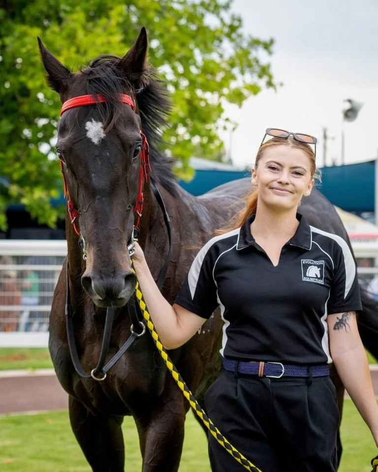 A young woman in a black and white polo shirt standing next to a dark horse with a red bridle and a white star on its forehead, holding the horse's lead rope with her left hand, outdoors in a green setting.