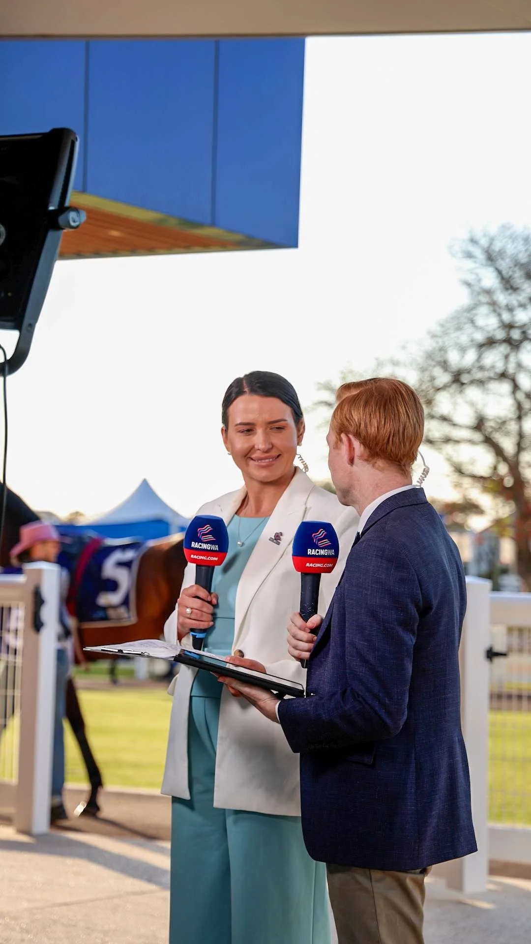 Two reporters with microphones labeled 'RACINGWA' are interviewing a woman at a horse racing event. The woman is holding a clipboard and smiling, while the man is standing beside her. Horses and a fenced race track are visible in the background, alon