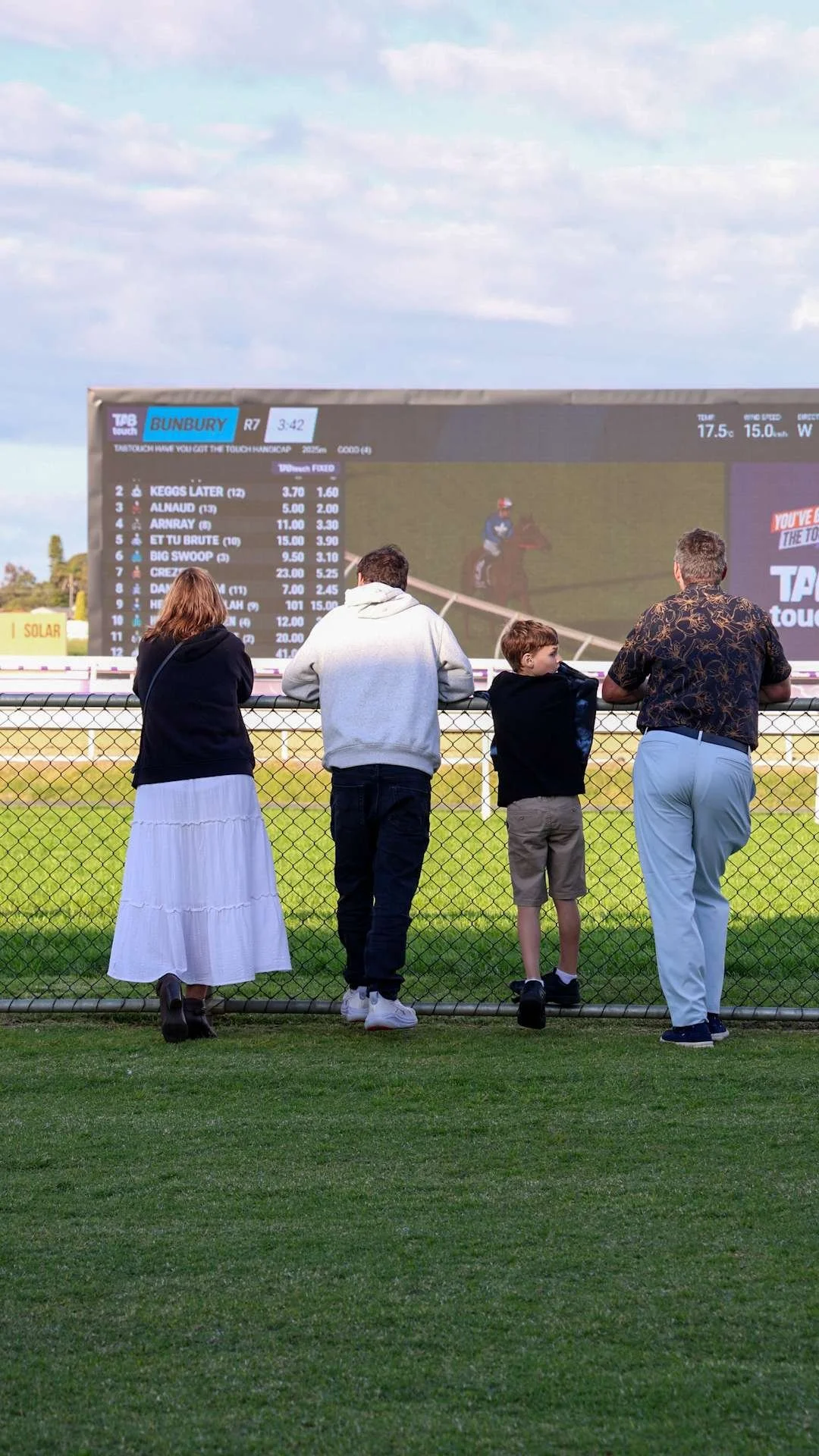 Four people standing behind a chain-link fence, watching horse racing on a large screen at BTC racetrack.