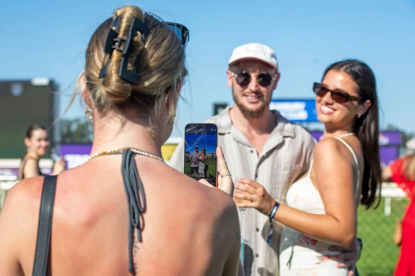 People taking a photo at an outdoor event on a sunny day, with a woman in a black dress in the foreground and a man and woman posing together in the background.