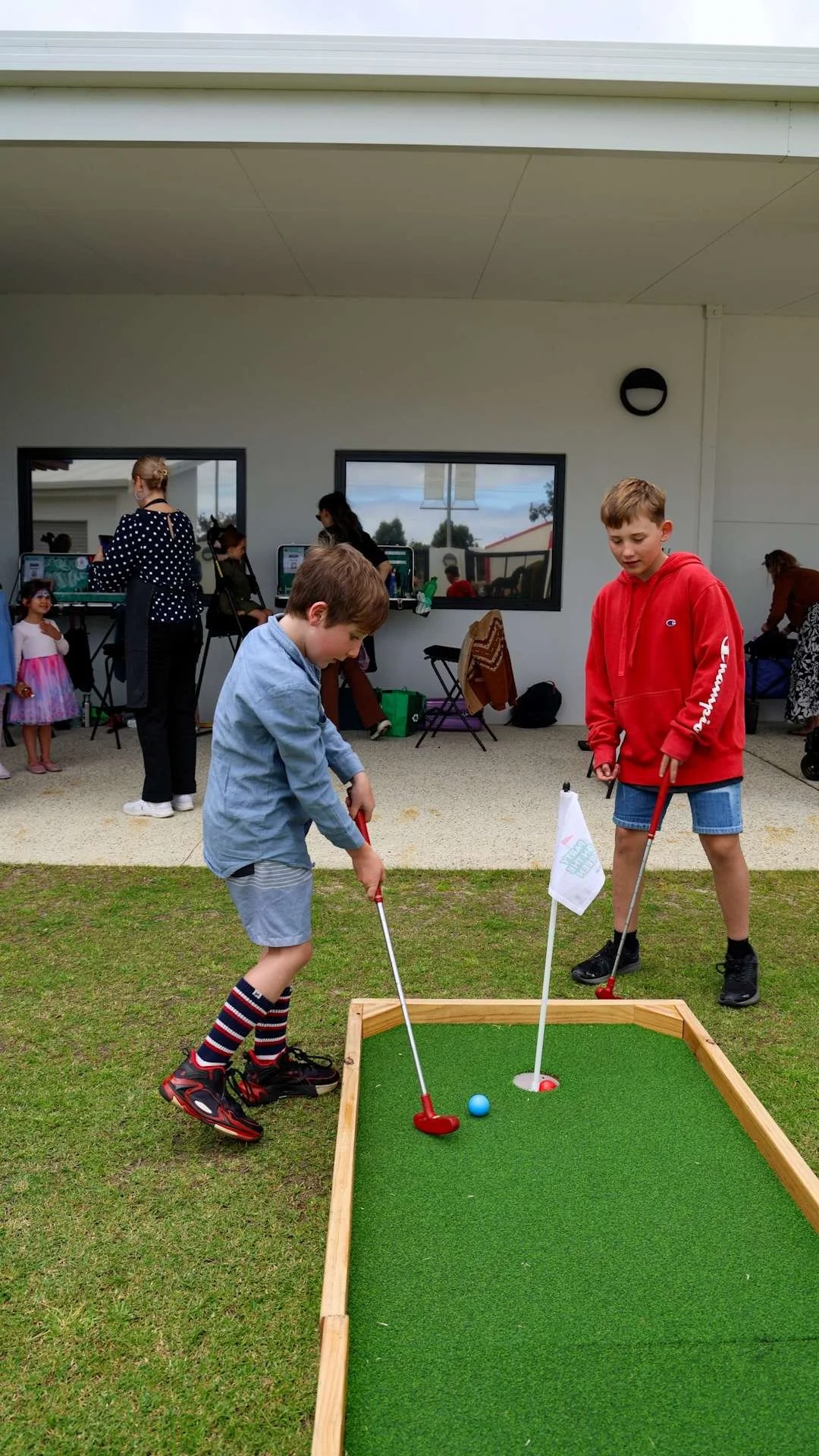 Two boys playing miniature golf outdoors on a small course, with a building and people in the background at Bunbury Turf Club Family Fun Day.