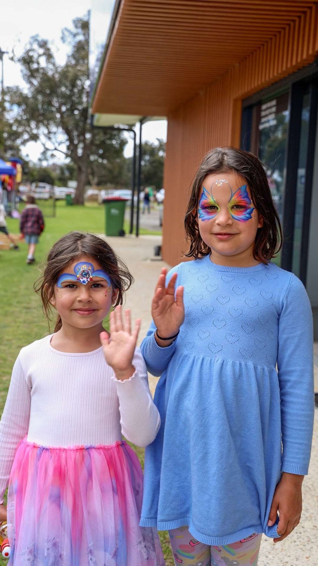 Two young girls standing outdoors, smiling and waving at the camera at Bunbury Turf Club Family Fun Day.