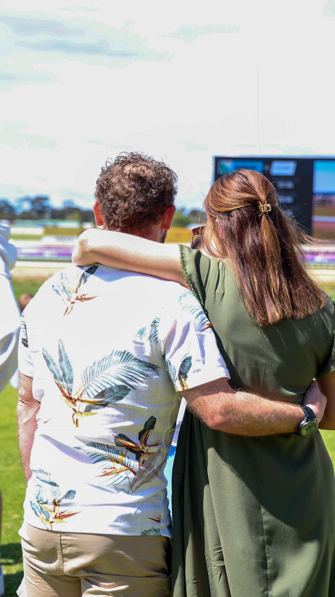 A couple is standing outdoors at a race track, with the woman resting her arm on the man's shoulder, watching a screen at the Bunbury Turf Club race course.