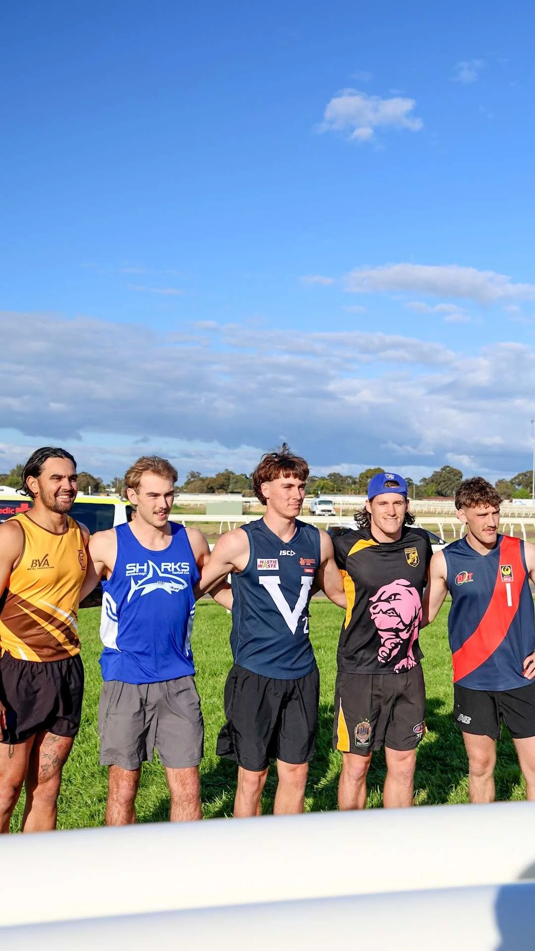 Five young men standing on a grassy field with their arms around each other, wearing athletic jerseys, suggesting a team before or after a sports event, under a partly cloudy sky.