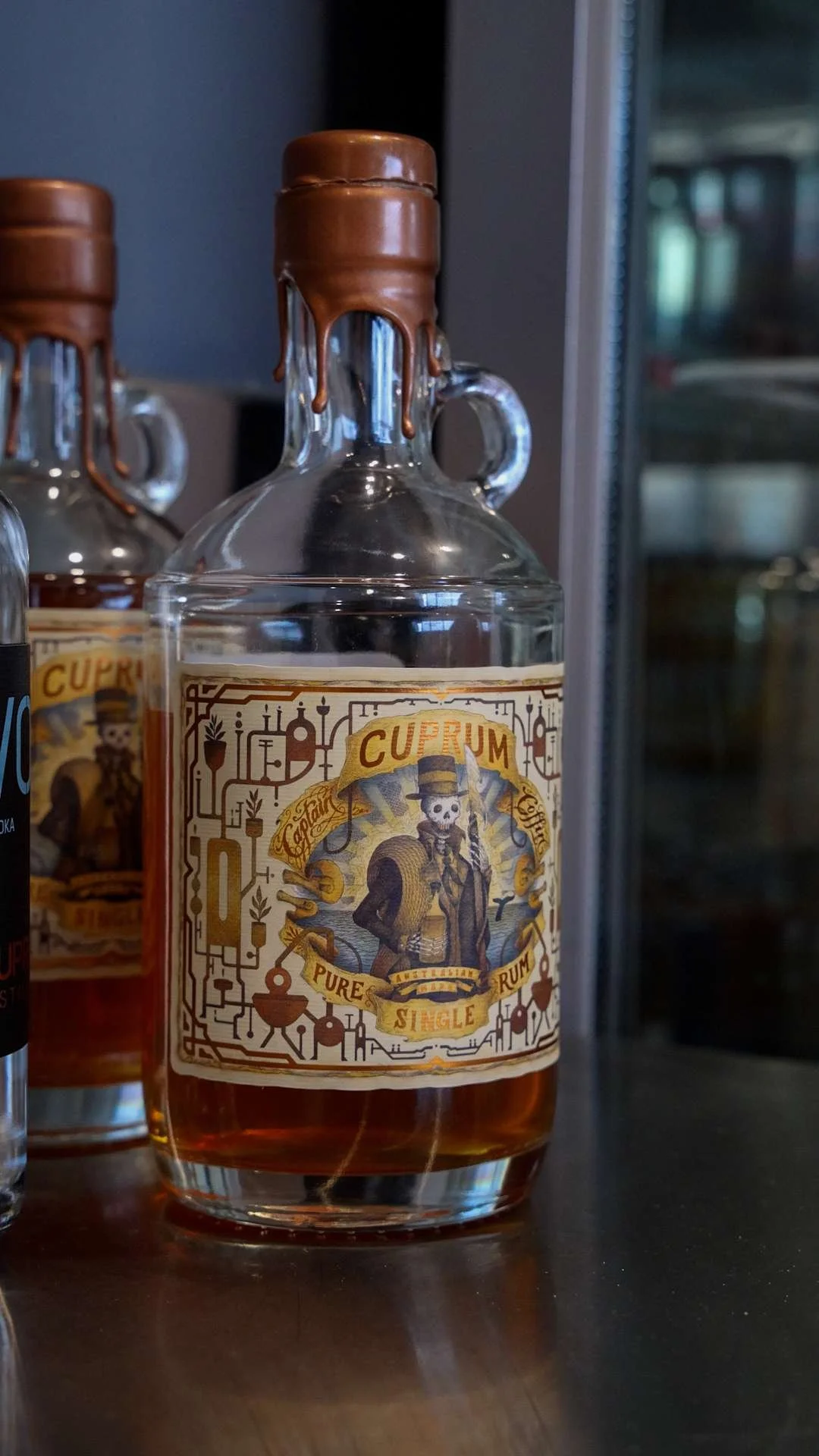 A clear glass bottle of Australian Cuprum Distillery rum with a label featuring a skeleton man in a hat and suit, holding a cane, and surrounded by decorative elements.