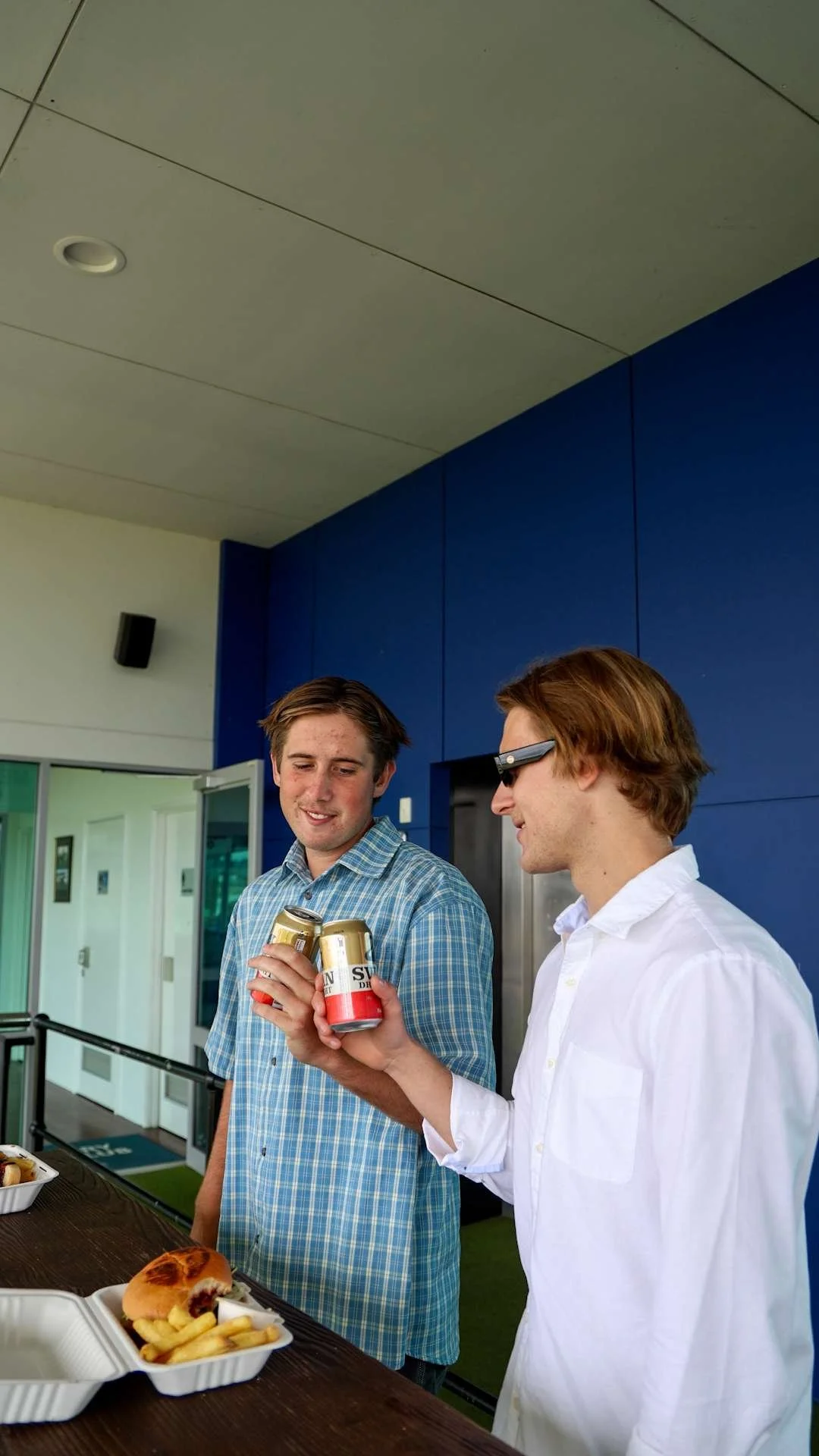 Two young men standing at an outdoor table on a race day, one holding cans of soda and the other looking at them.