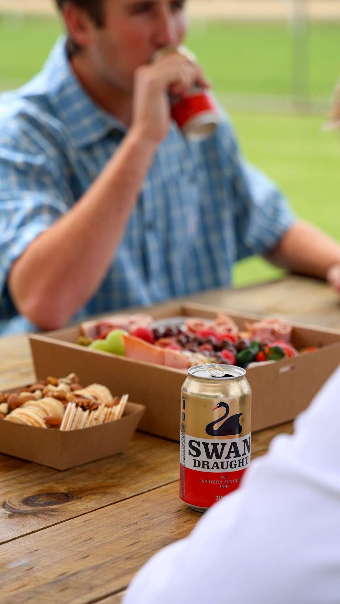 A person drinking from a red can, with a picnic table that has snacks, a box of fruit, and a can of Swan Draught beer, outdoors on a sunny day.