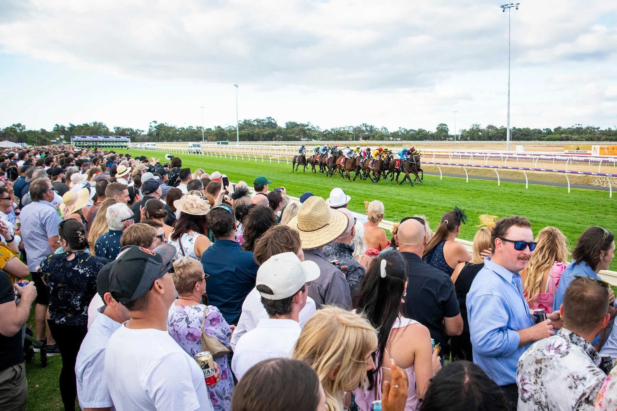 Crowd of spectators watching horse race on a green racetrack with multiple horses and jockeys in colorful uniforms, under partly cloudy sky.