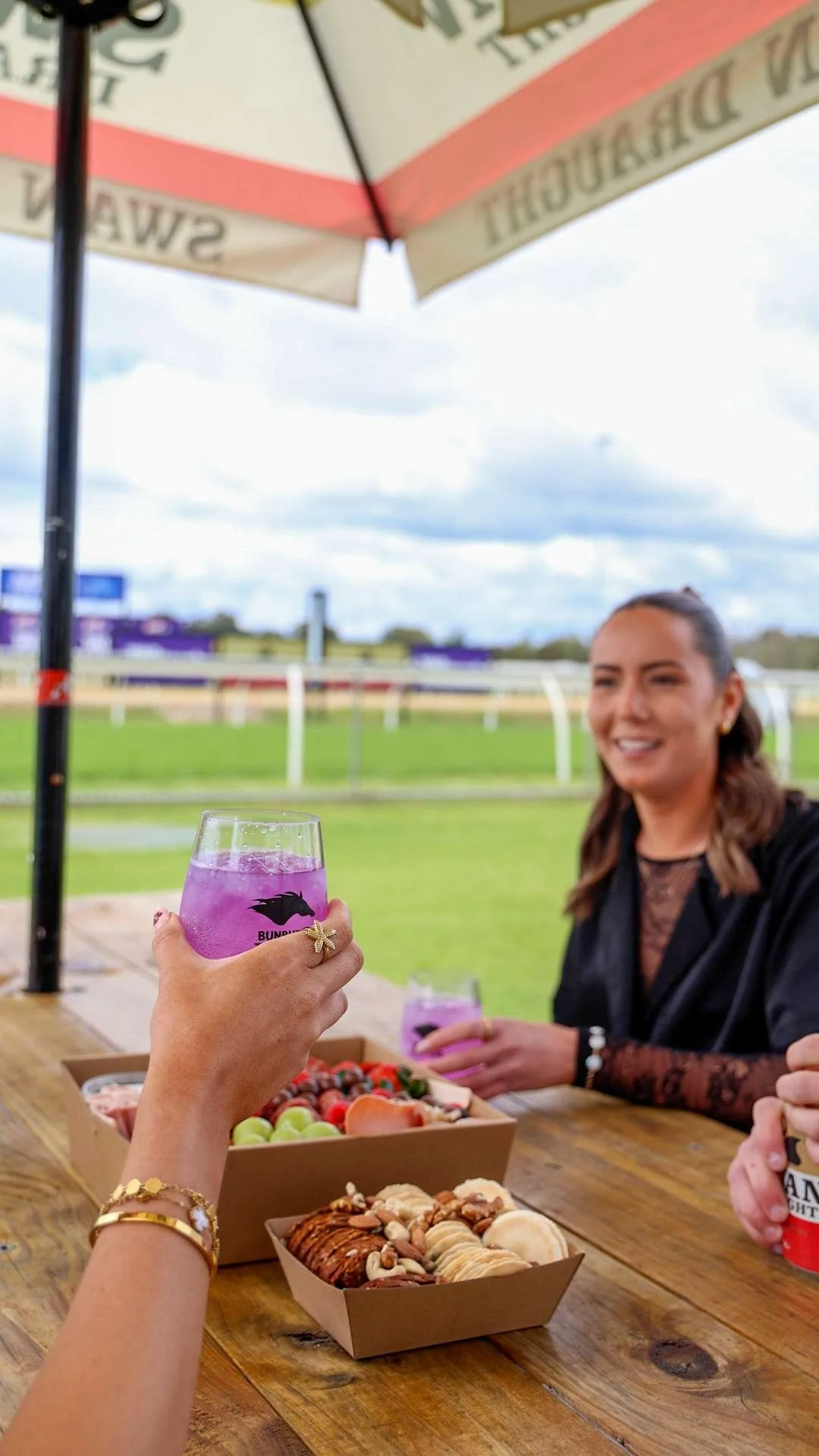 People sitting at a wooden table under a large umbrella, with a view of a racetrack in the background. One person is holding a pink drink in a stemmed glass, and another person is smiling. There are boxes of assorted cookies and fresh fruit on the table.