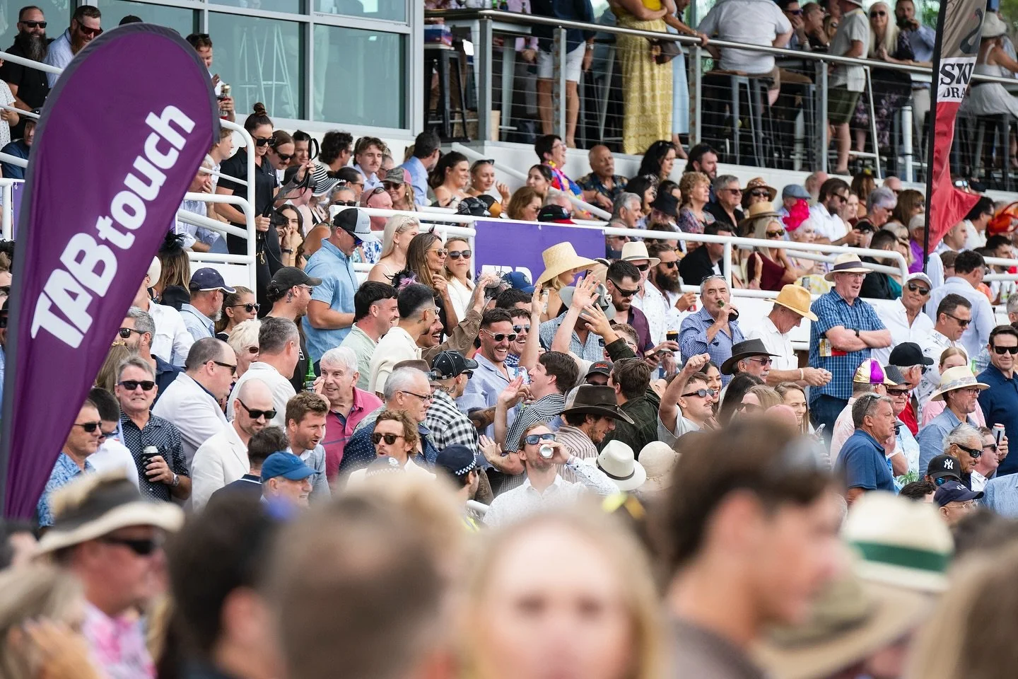 Crowd of people at an outdoor event, many wearing sunglasses and hats, in attendance at a sports or entertainment event.