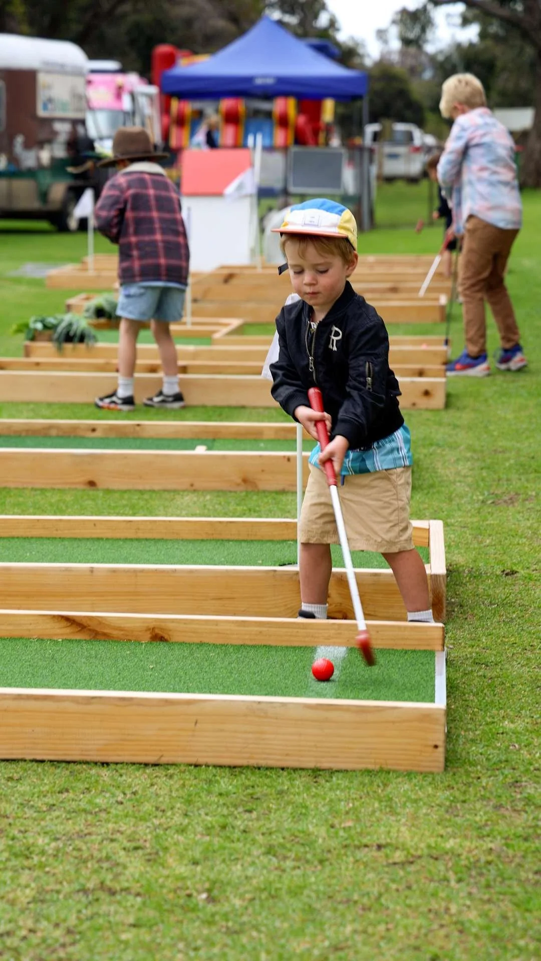 Kids playing mini-golf outdoors, a young boy in a baseball cap and black jacket putting a red golf ball on green artificial turf in wooden-framed mini-golf course, with other children in the background and a food truck and game tents.