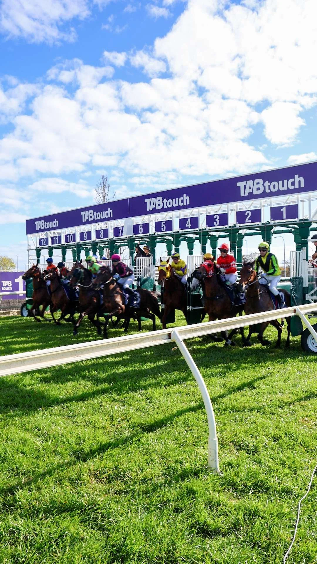 Horse race start with multiple horses and jockeys in colorful uniforms, lined up on the racetrack under a bright blue sky with scattered clouds.
