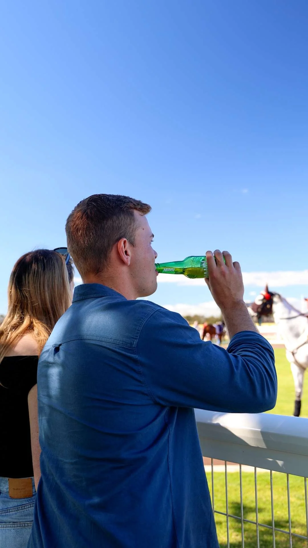 A man with short hair in a blue shirt drinking from a green glass bottle at a horse racing event on a sunny day.