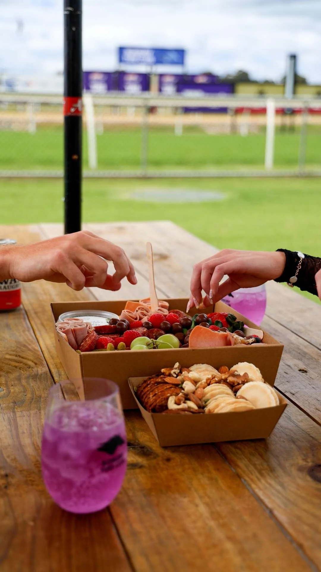 A picnic table with a cardboard box of assorted fruit, snacks, and chocolates, and two hands reaching into the box, with a sports field and purple billboard in the background.
