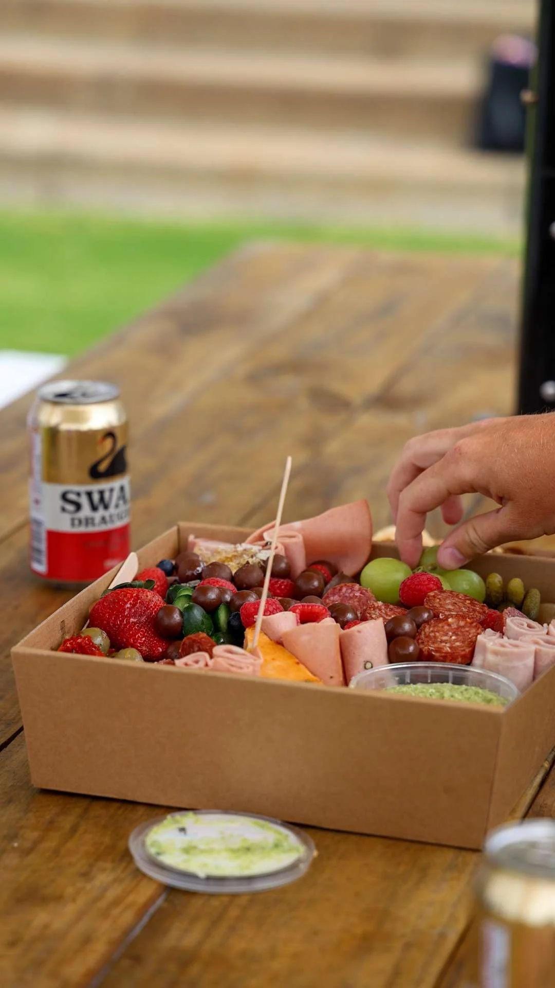 A hand reaching for food in a box of assorted meats and fruits, with a can of Swamp Draft beer on a wooden table outdoors, blurred background of steps and grass.