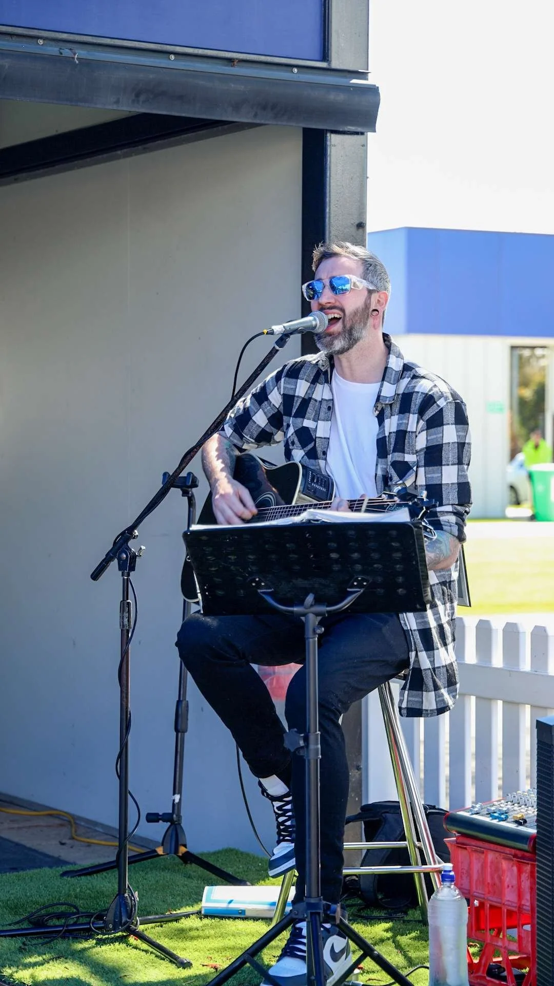 A man wearing sunglasses and a checkered shirt, singing into a microphone while playing an acoustic guitar outdoors at Bunbury Turf Club Family Fun Day.