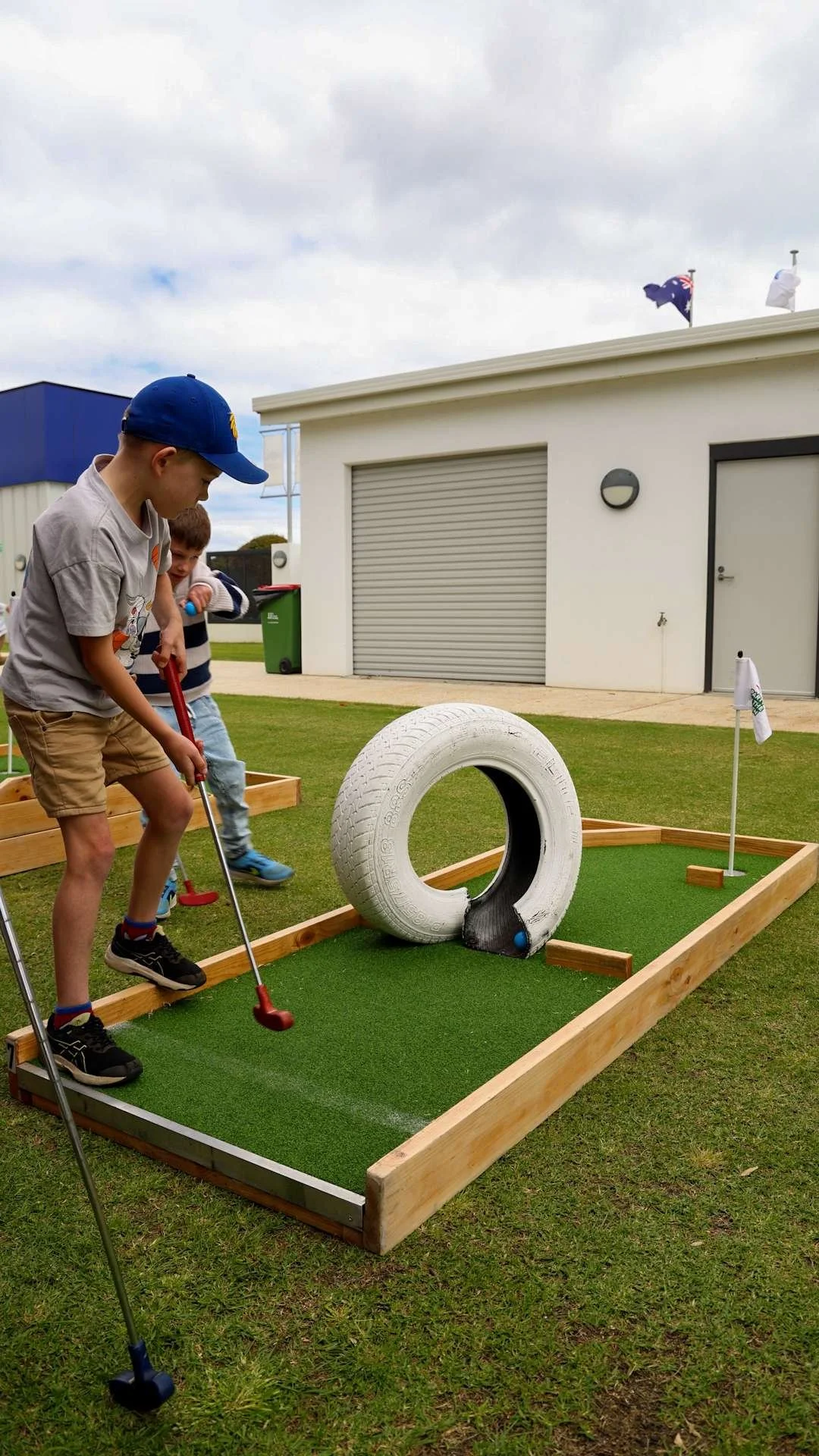 Two boys playing miniature golf on a small green course outdoors with a tire obstacle and hole with flag at Bunbury Turf Club Family Fun Day.