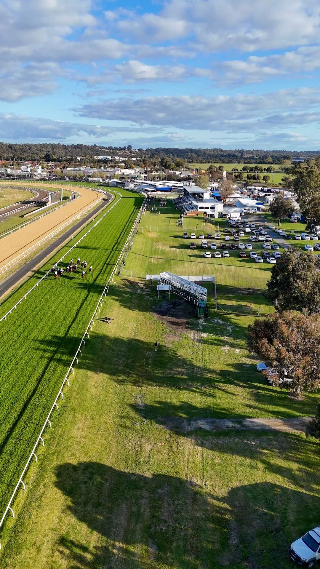Aerial view of a horse racing track with green grass, racing lanes, and horses with jockeys riding them, alongside parking lots filled with cars and buildings, under a partly cloudy sky.