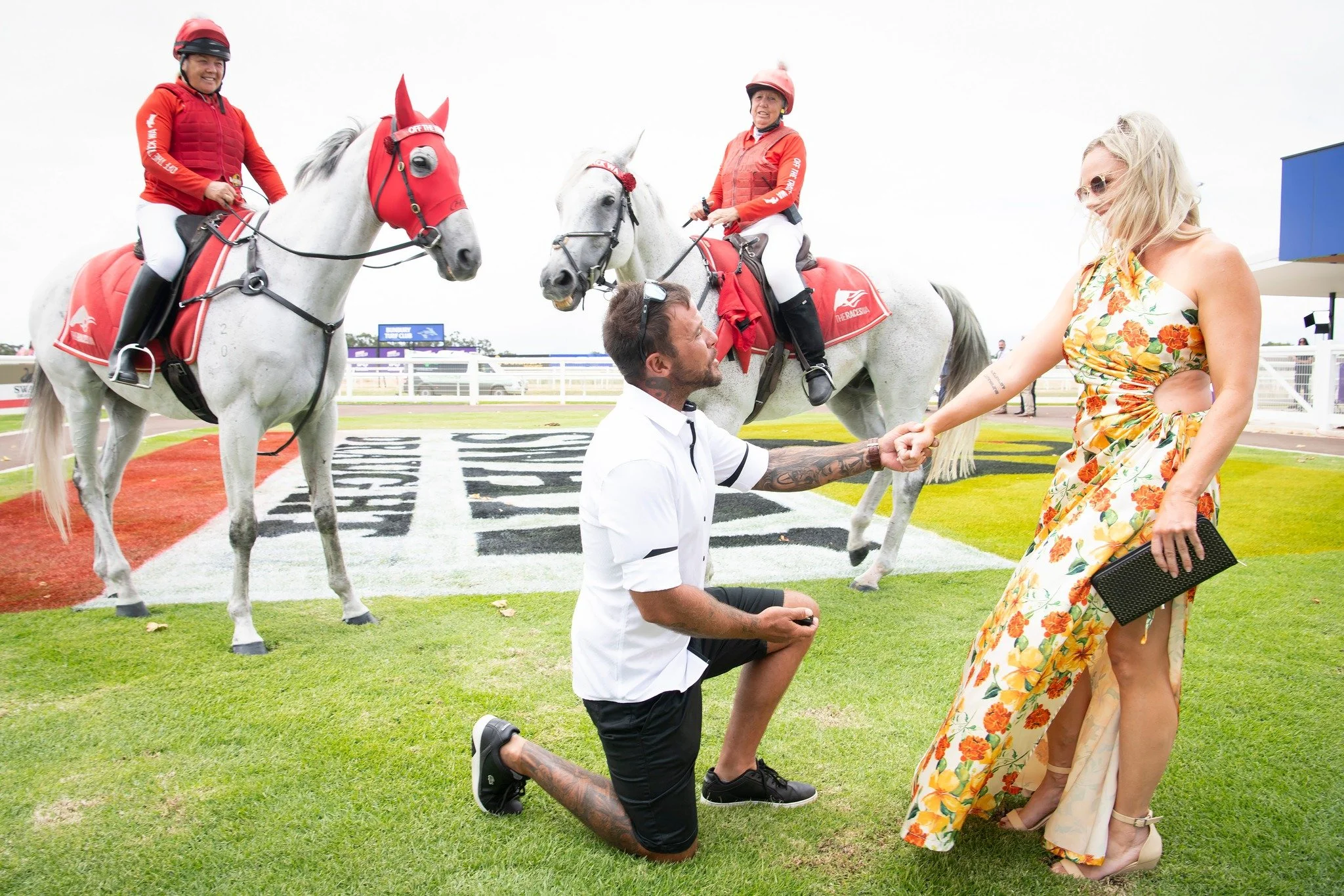 A man kneeling on one knee holding hands with a woman at a horse racing event. Two women are on horseback in the background, dressed in racing gear with red saddle blankets. The setting is a racecourse with grass, fencing, and a cloudy sky.