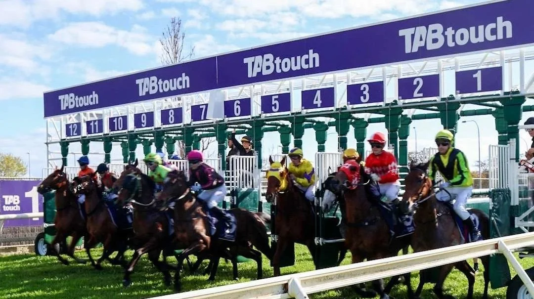 Horse racetrack with three jockeys riding horses and a crowd of spectators behind a fence, under a clear blue sky.