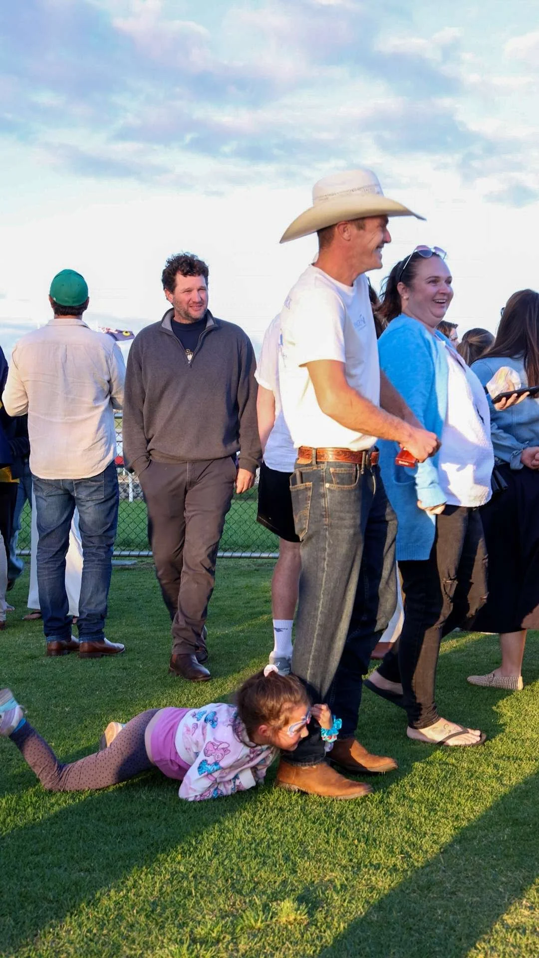 A girl lies on the grass, holding onto a man's leg, at a racing event on a grassy field with a chain-link fence and cloudy sky in the background.