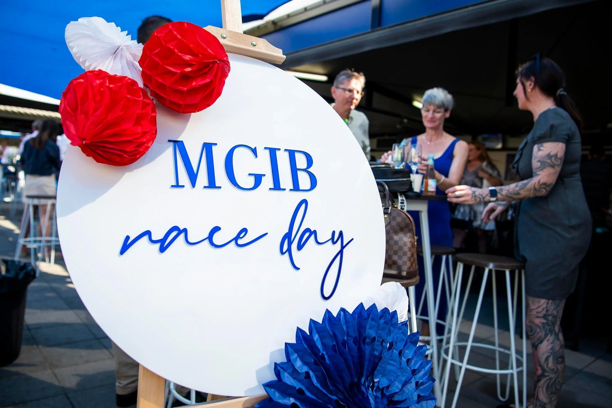 A sign with blue text that reads 'MGIB race day' decorated with red and blue paper flowers, at an outdoor event with people and a bar in the background.