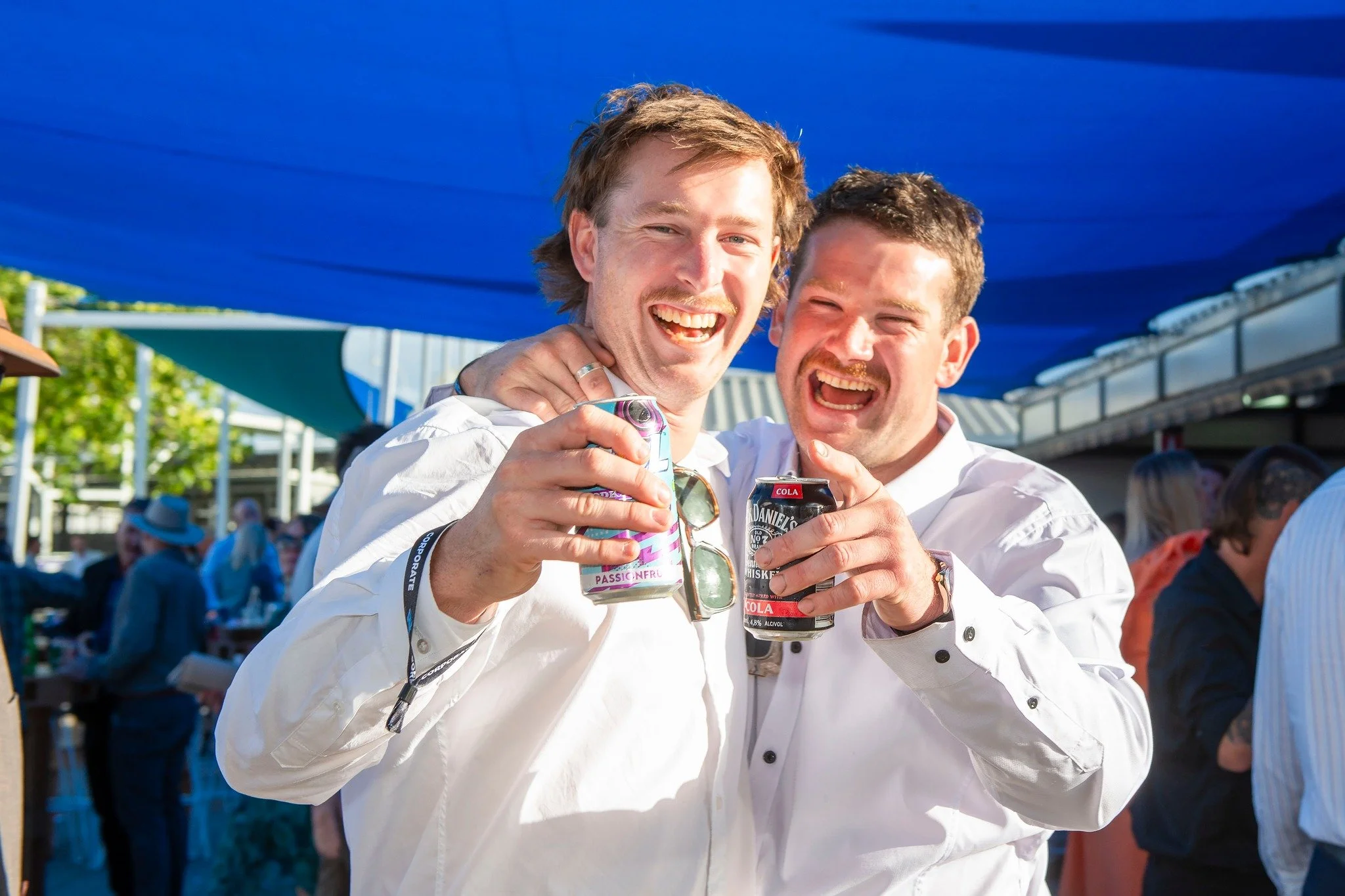 Two men smiling and holding soda cans at an outdoor event under a blue canopy.