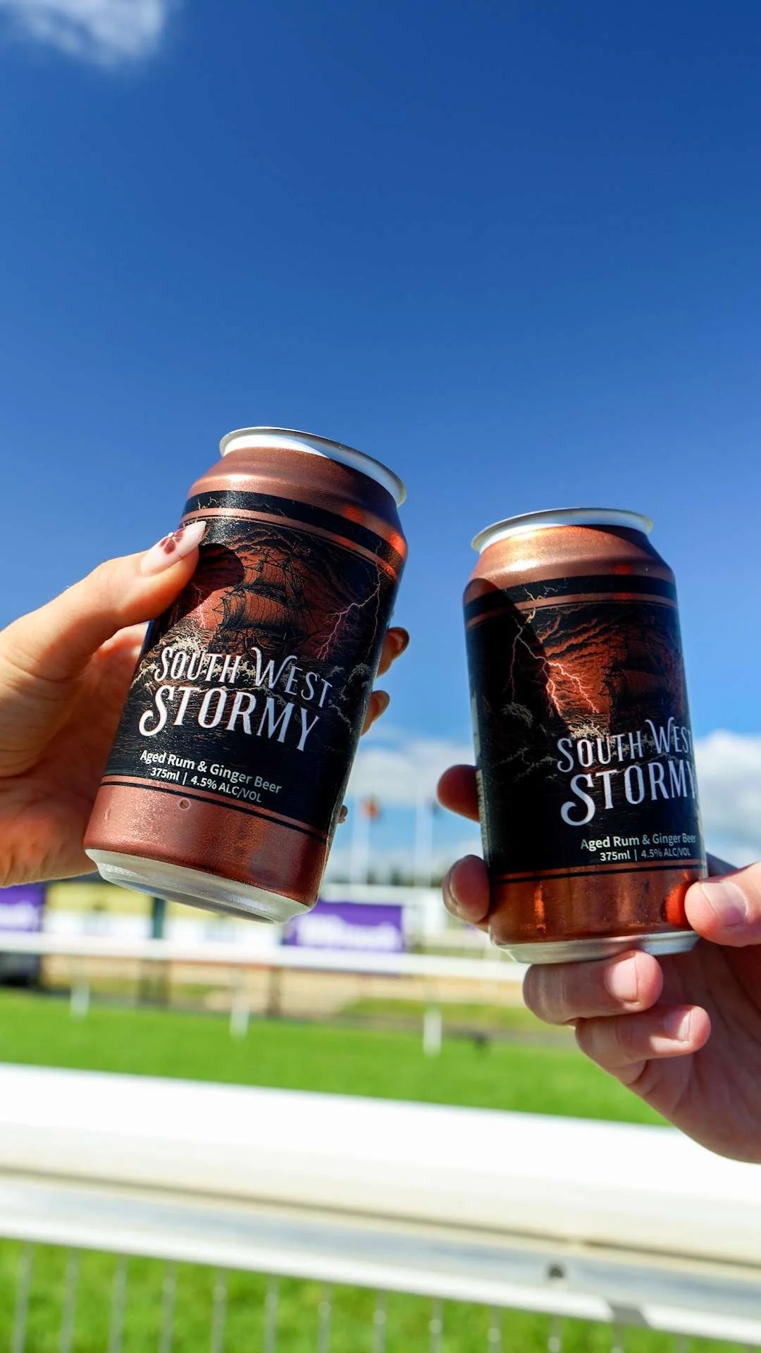 Two cans of South West Stormy aged rum and ginger beer held up against a clear blue sky at BTC racetrack