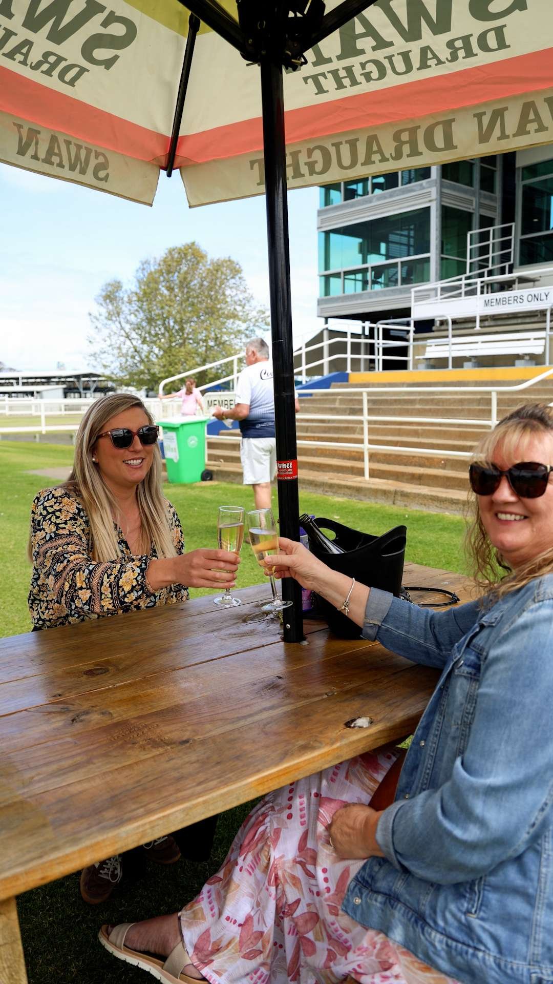 Two women sitting under a large beach umbrella at a wooden picnic table, clinking glasses of champagne or sparkling wine at Bunbury Turf Club racecourse.