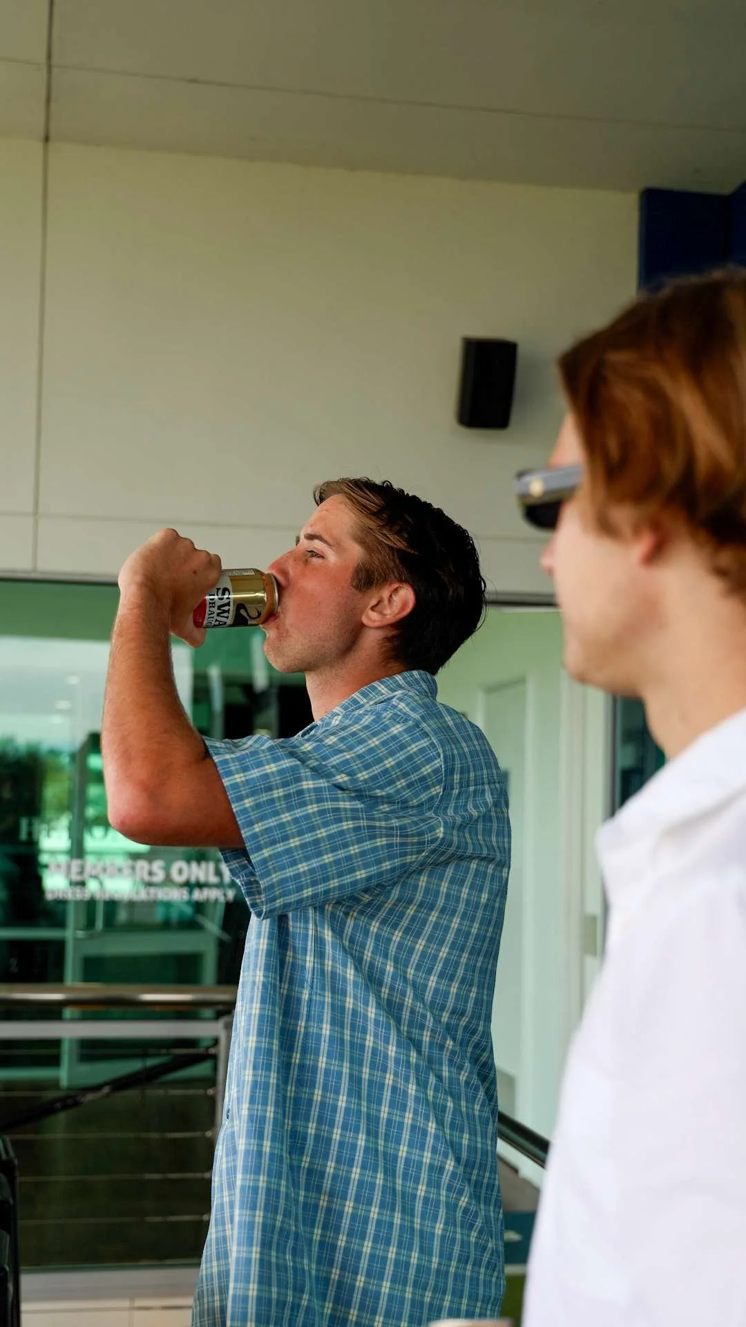 A man in a blue plaid shirt is drinking from a can while another man, wearing glasses, watches him.