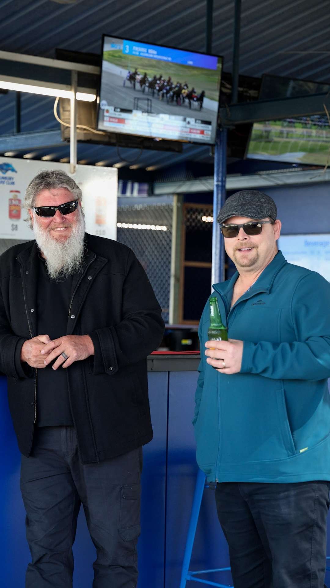 Two men wearing sunglasses, one with a beard and black coat, the other in a gray hat and blue jacket, standing indoors near a bar with screens displaying horse racing in the background, holding beverages at Bunbury Turf Club.