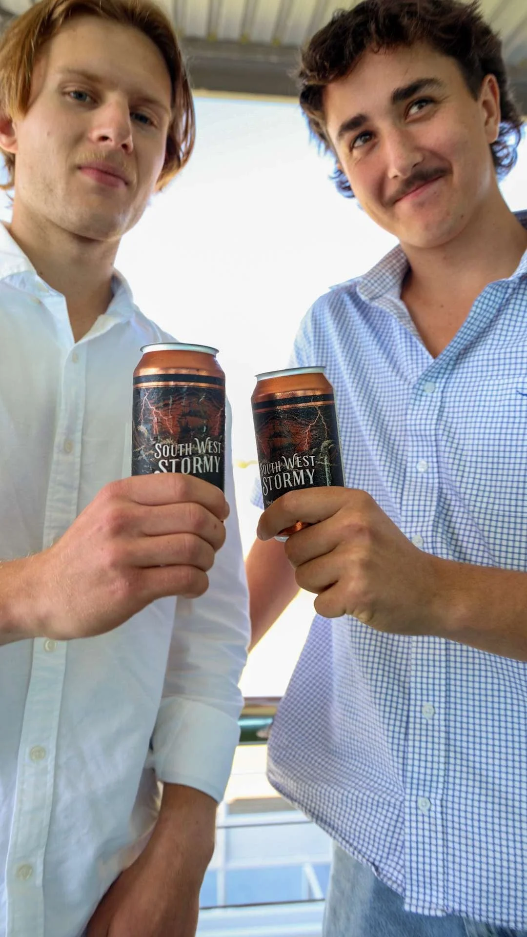 Two young men in button-up shirts holding cans of South West Stormy beer, smiling and standing outdoors at Bunbury Turf Club.