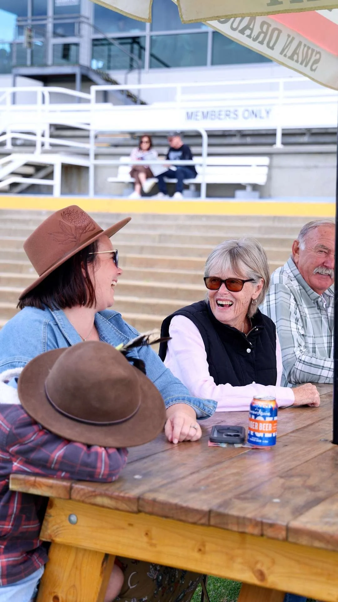 Four people sitting at a wooden outdoor table, smiling and chatting. Two women and two men, some wearing sunglasses, with a can of ginger beer and a phone on the table enjoying a day out at Bunbury Turf Club.