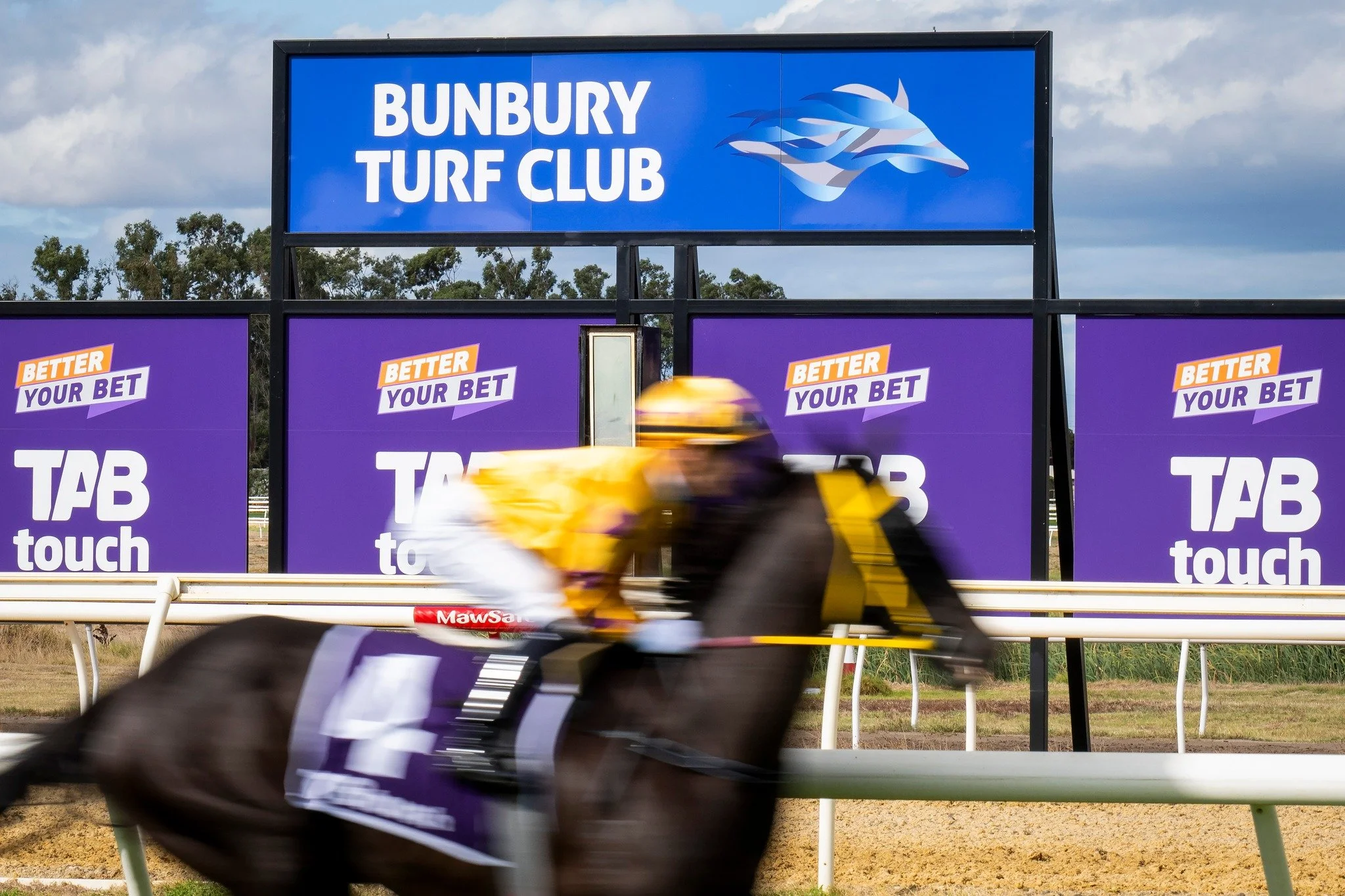 A horse race at Bunbury Turf Club with a jockey in yellow and black riding a dark horse, blurred from speed, with a digital advertising billboard in the background displaying the words 'Bunbury Turf Club' and various betting advertisements.