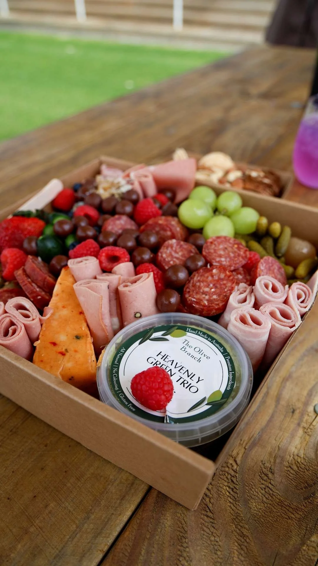 A wooden box filled with a variety of meats, cheeses, and fruit, with a small container labeled "Heavenly Green Trio" in the foreground, set on a wooden table outdoors at Bunbury Turf Club Family Fun Day.