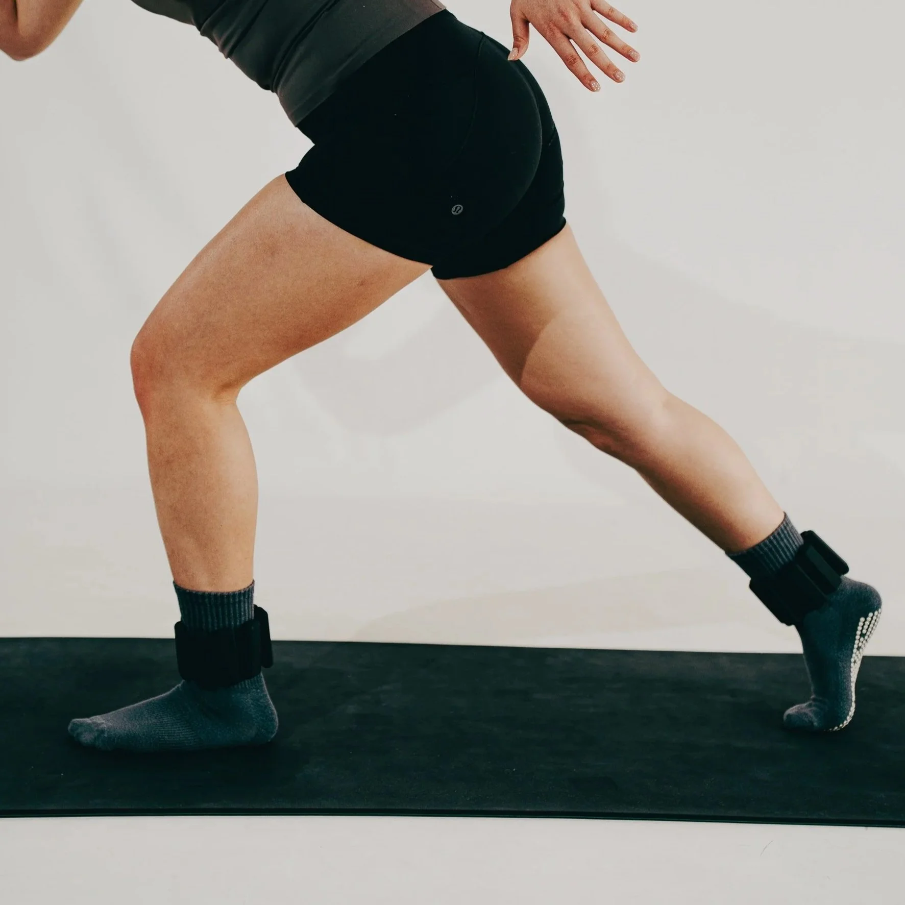 Person stretching in a lunge position on a black exercise mat, wearing black shorts, socks, and an ankle weight on each leg.