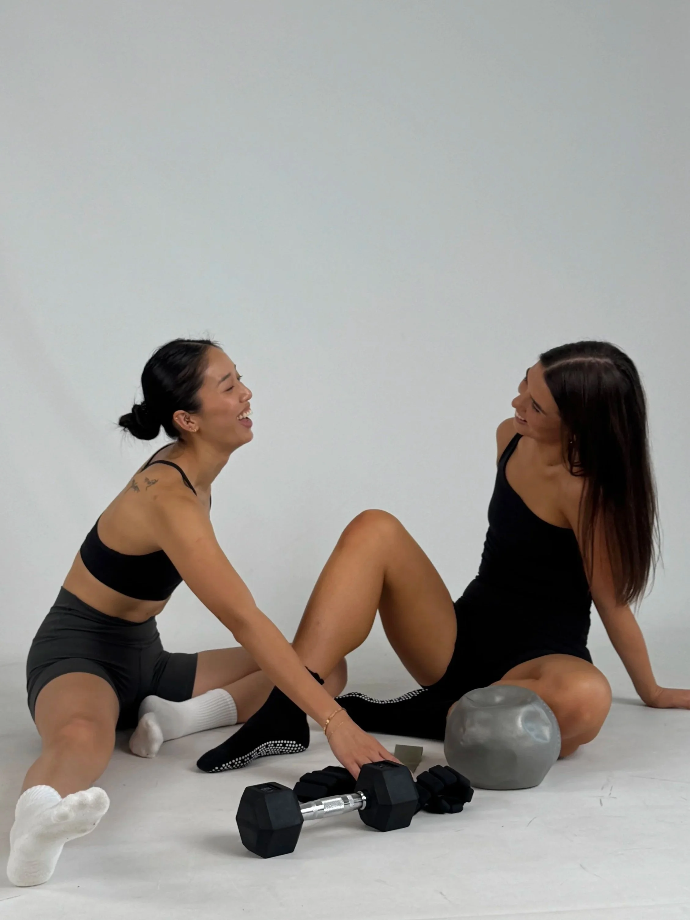 Two women sitting on the floor, laughing, with weightlifting equipment including dumbbells, ankle weights, and a pilates ball around them.