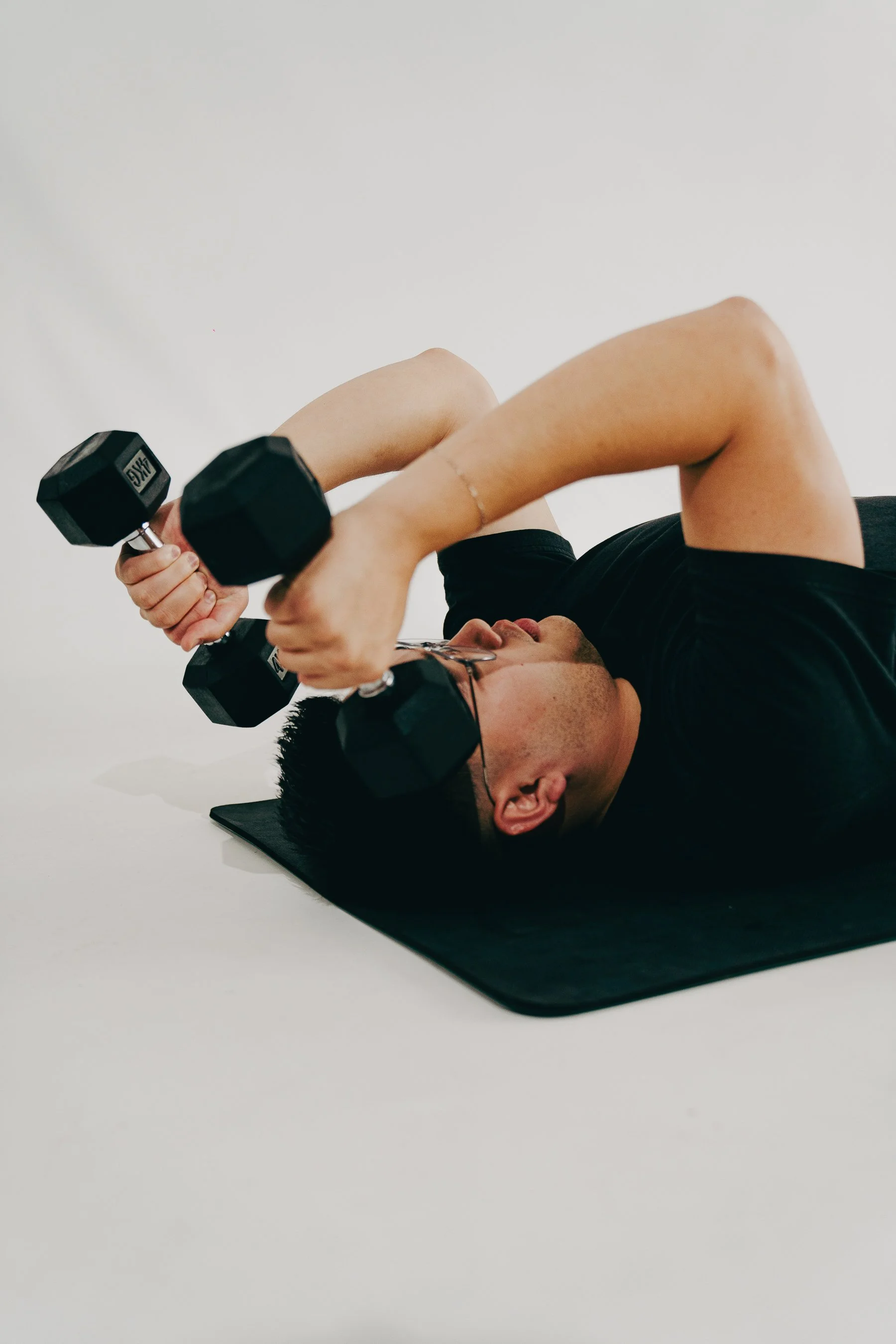 Man lying on a workout mat lifting dumbbells in a boutique studio setting.