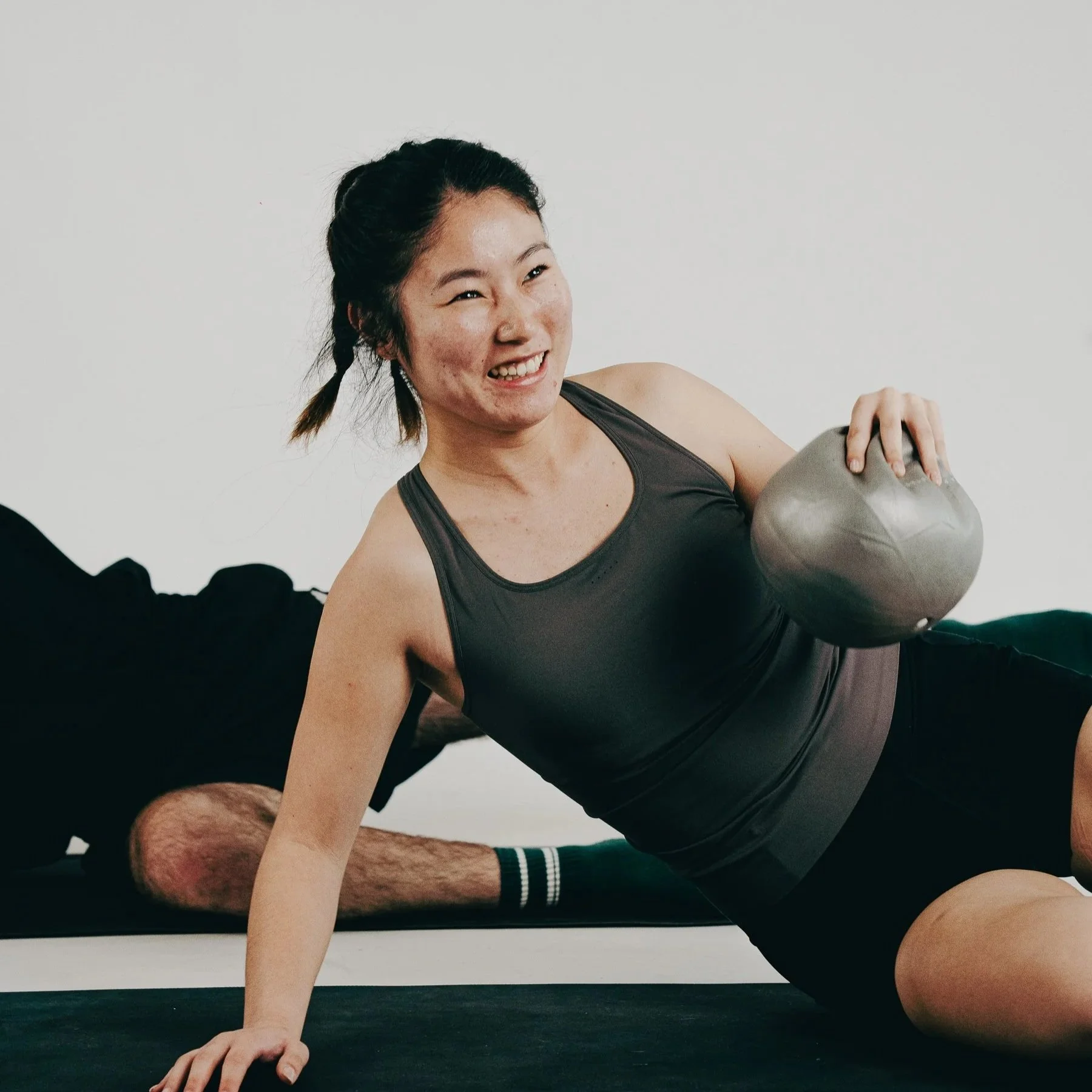 A woman with a ponytail smiling while doing a side plank exercise with a ball in a boutique pilates studio setting.