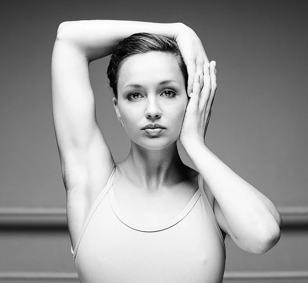 Black and white portrait of a young woman with short hair, wearing a sleeveless top, with one arm raised and hand resting on the side of her face, looking directly at the camera.