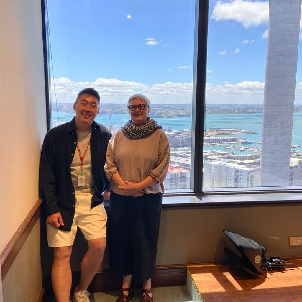 Two people stand inside a room with a large window showing a city skyline and water outside. One is a young man, and the other is an older woman, both smiling at the camera.
